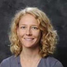 lady with blonde curly hair wearing grey shirt and necklace in front of grey background